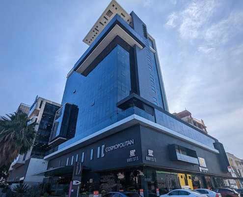 Tall blue-glass mixed-use building with Cosmopolitan signage and storefronts at street level, cars parked outside