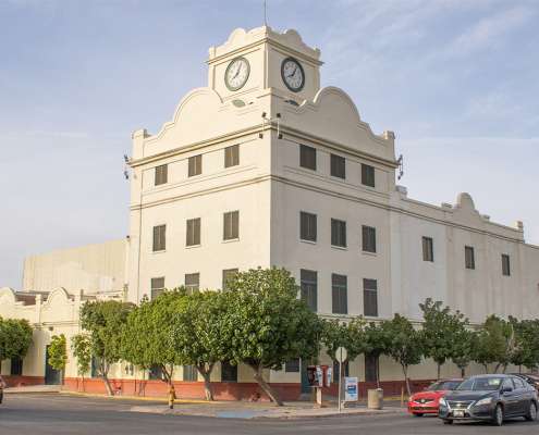 White multi-story building with a central clock tower, trees in front, and cars parked along the street.