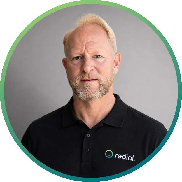 Portrait of a middle-aged man with blond hair and a beard, wearing a black polo shirt with a redial logo against a gray studio background.